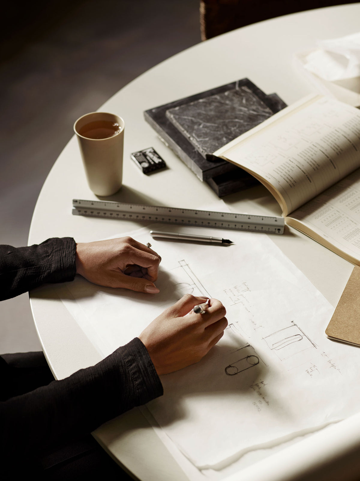 Hands sketching technical drawings on white paper at a round table with ruler, pen, open book and cup of tea