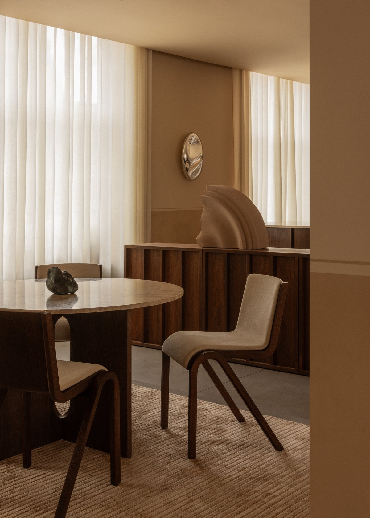 Minimalist dining area with rounded wooden table, curved beige chairs, sheer white curtains and sculptural decor