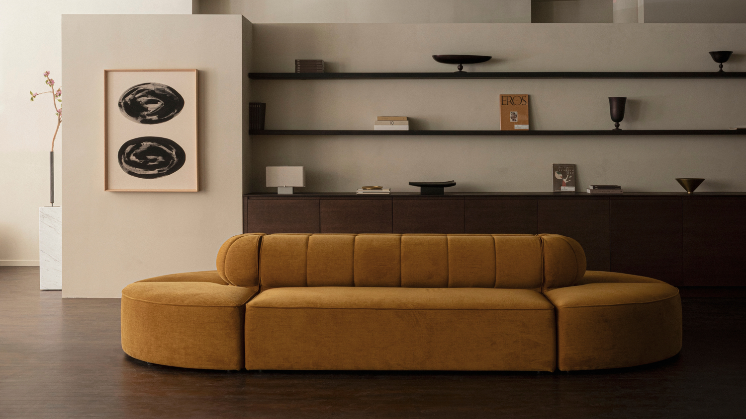 Mustard velvet curved sofa with channel-tufted back in minimalist living room with dark shelving and abstract wall art