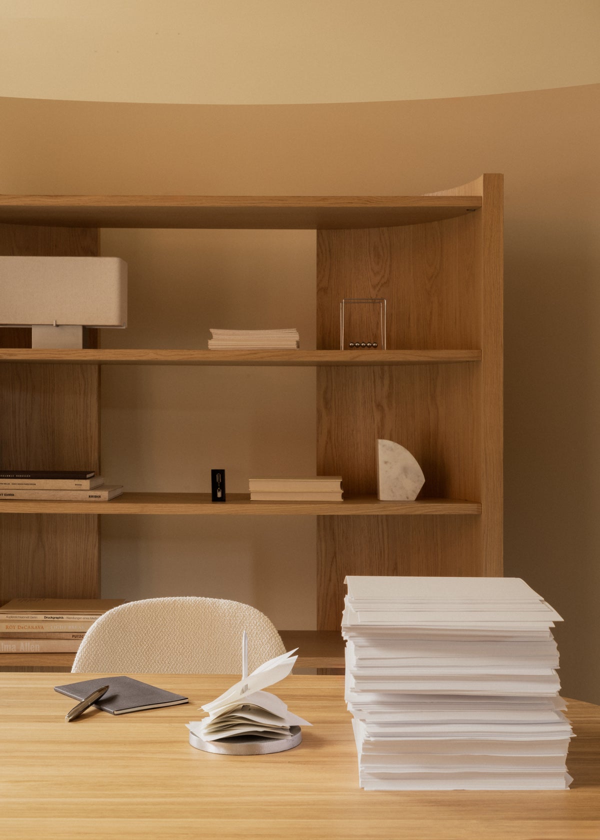 Minimal wooden desk and shelving with stacked white papers, notebook, pen and simple decorative objects