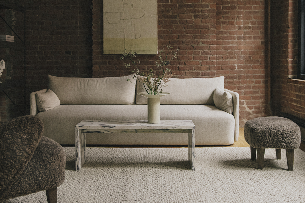 Neutral-toned living room with a beige sofa, wooden coffee table holding a vase of branches, textured rug and brick wall backdrop