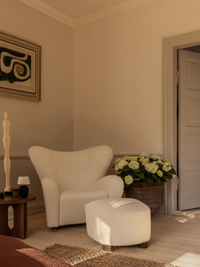 The Tired Man Ottoman in white bouclé, paired with the matching armchair in a sunlit corner next to a wooden side table.