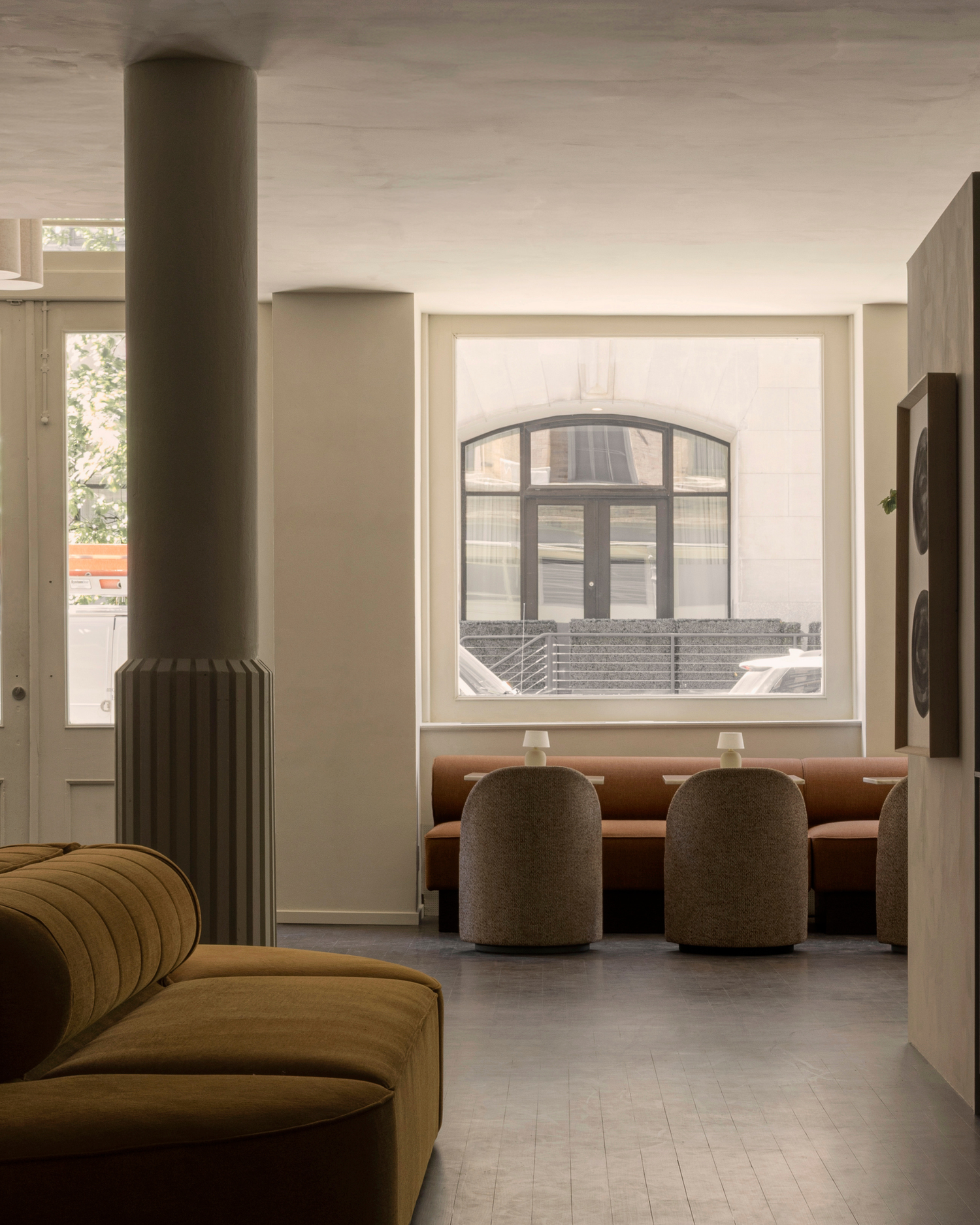 Minimal interior with tan upholstered sofa, rounded beige dining stools and orange banquette under large square window.
