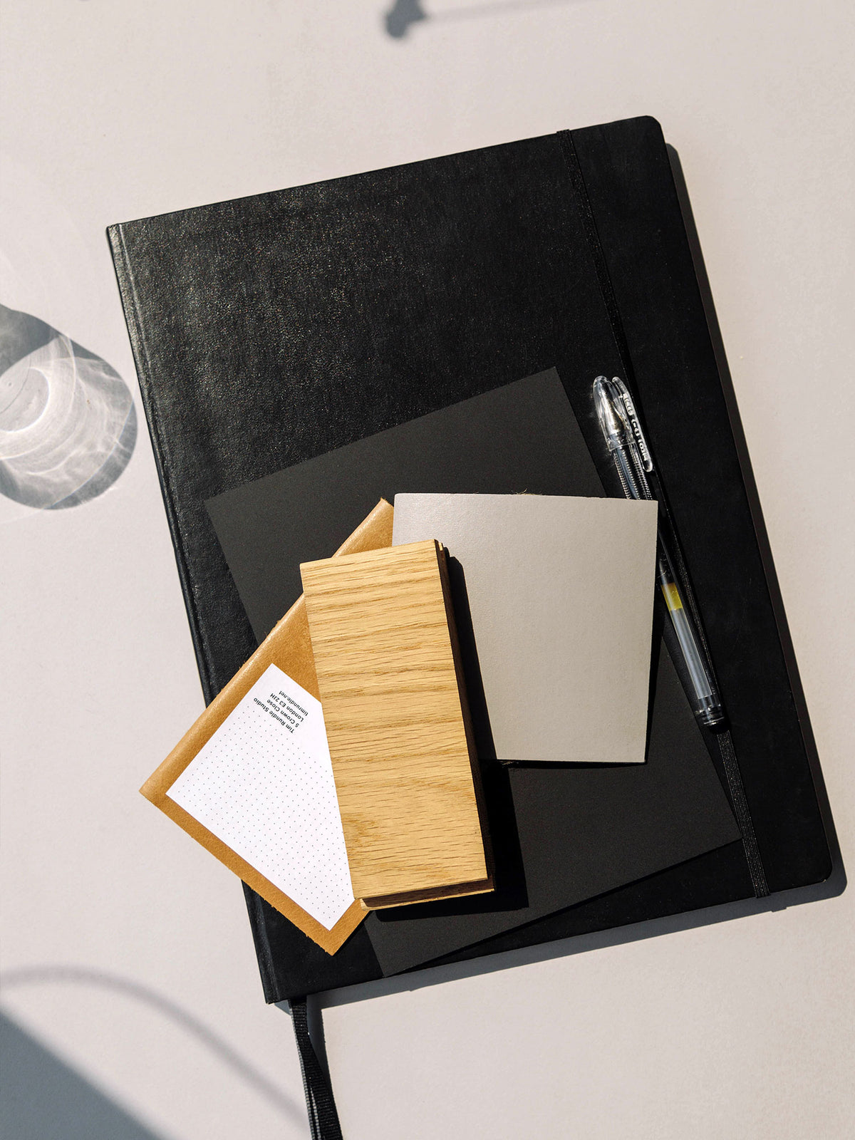 Top-down view of stacked black notebook, sample swatches, wooden block, envelope and pen on pale background