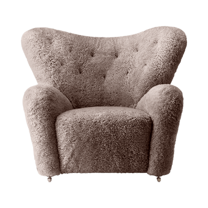 The Tired Man Lounge Chair in beige sheepskin with a tufted backrest and wooden legs with brass casters.