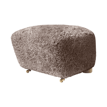 The Tired Man Ottoman upholstered in soft textured sheepskin in a neutral brownish shade, standing on small brass castors.