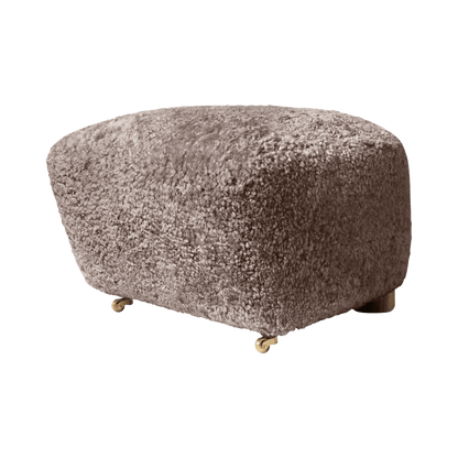 The Tired Man Ottoman upholstered in dark sheepskin, featured on small brass castor wheels against a white background.