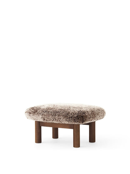 The Brasilia Ottoman, shown at an angle, with a softly curved cushion in Root sheepskin and a dark stained oak base.