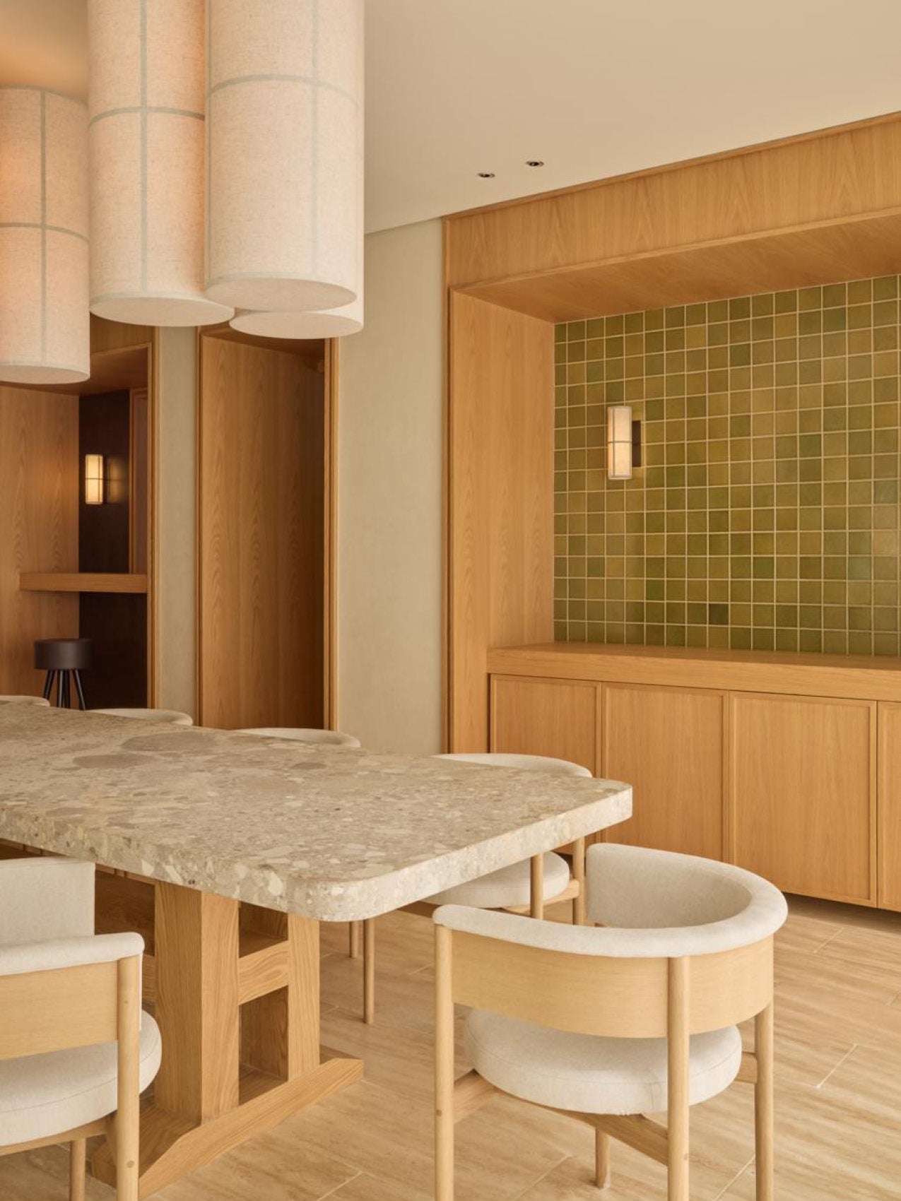 Dining space with terrazzo tabletop, light oak base and curved upholstered chairs beneath cylindrical fabric pendant lamps.