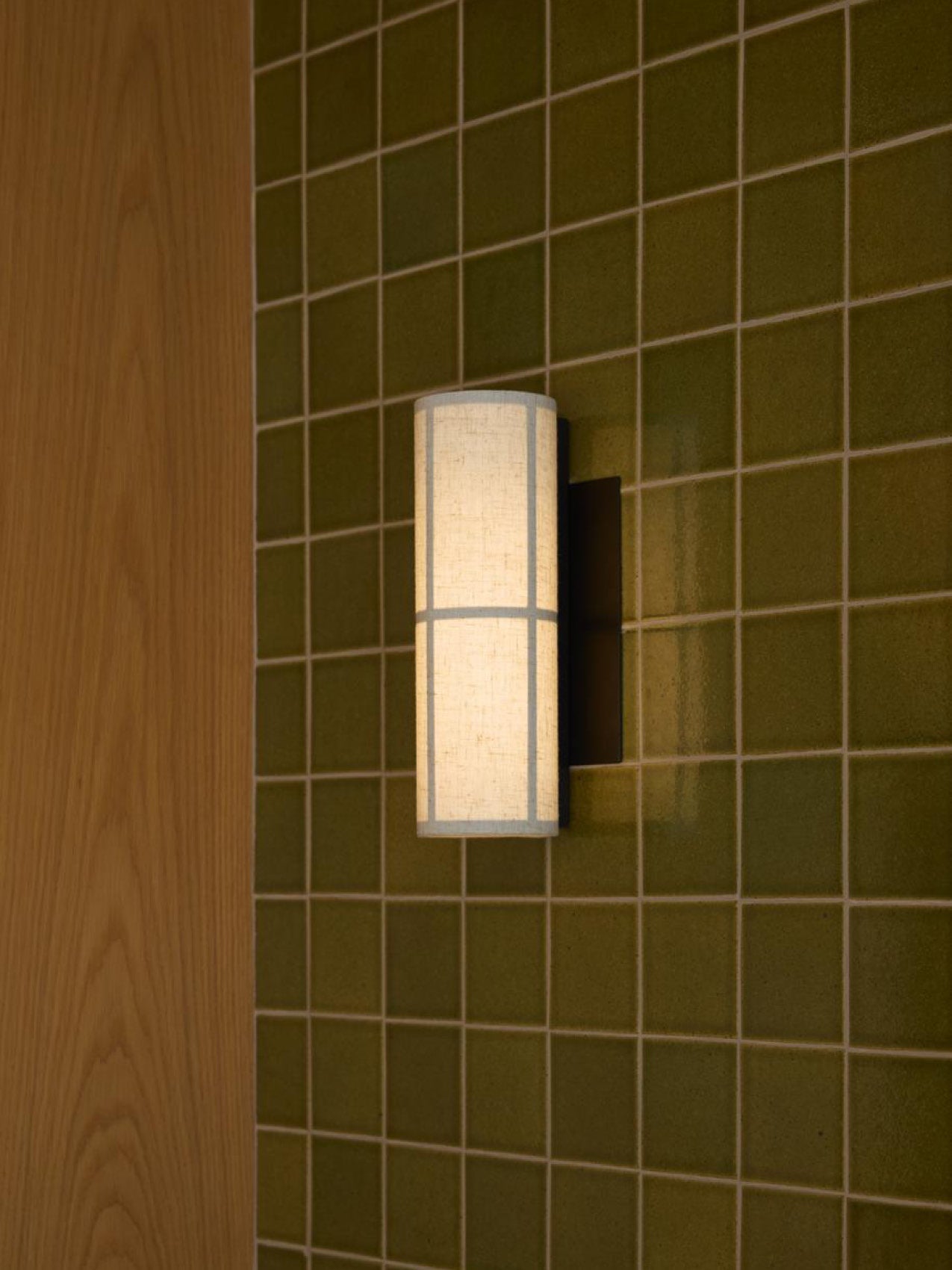 Cylindrical fabric wall sconce with grid seams, warm white glow on olive green square tiles and light wood panel.