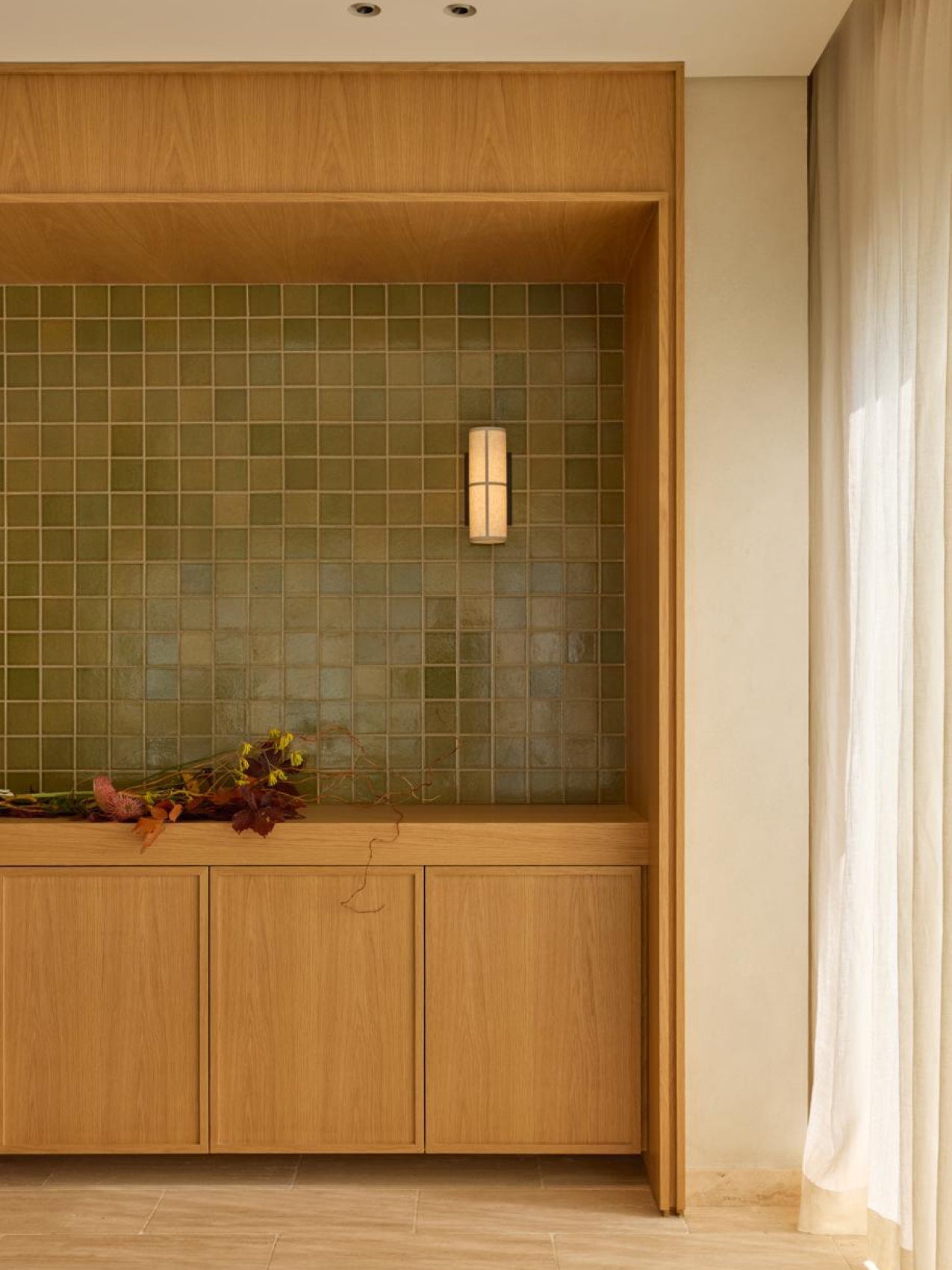 Oak cabinet with recessed niche of green square tiles, slim cylindrical wall sconce and dried floral arrangement on top