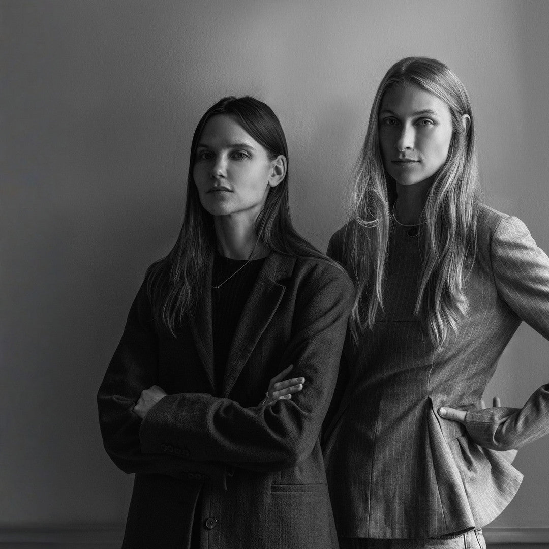 Two women wearing tailored blazers in dark and light gray wool, standing side by side against a plain wall.