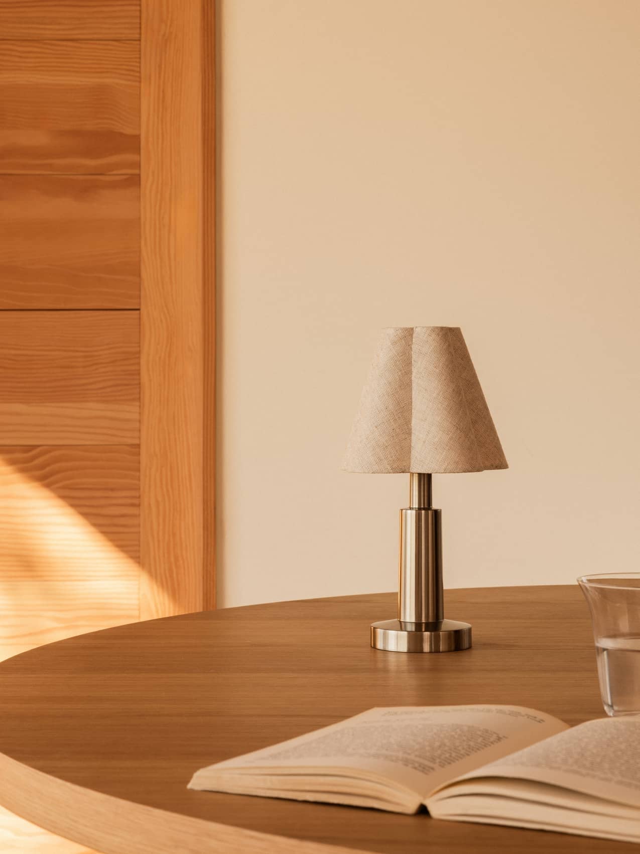 Admiral Portable Lamp with a sculpted fabric shade and metallic base sits on a wooden dining table beside an open book.