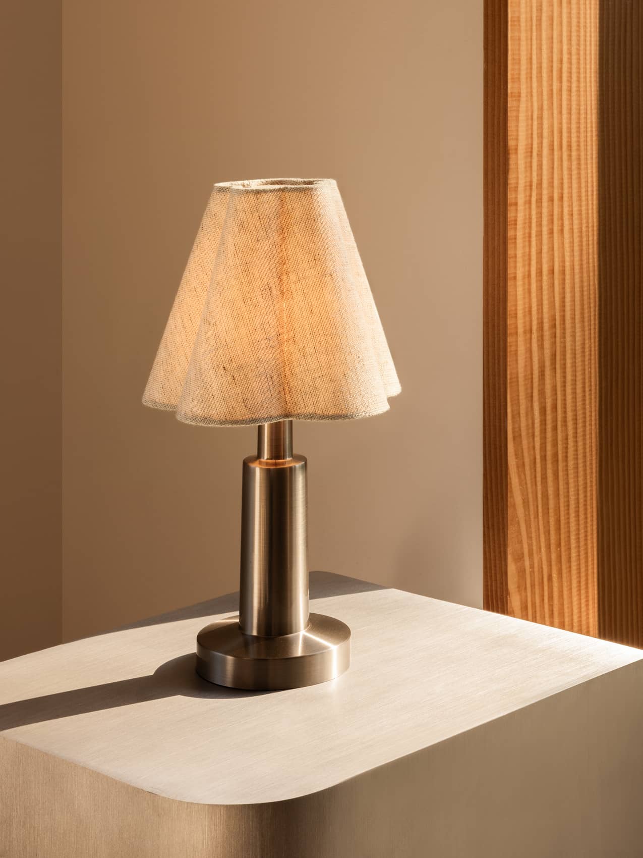 Admiral Portable Lamp with a sculpted fabric shade and brushed metal base, glowing on a light-colored wooden table.