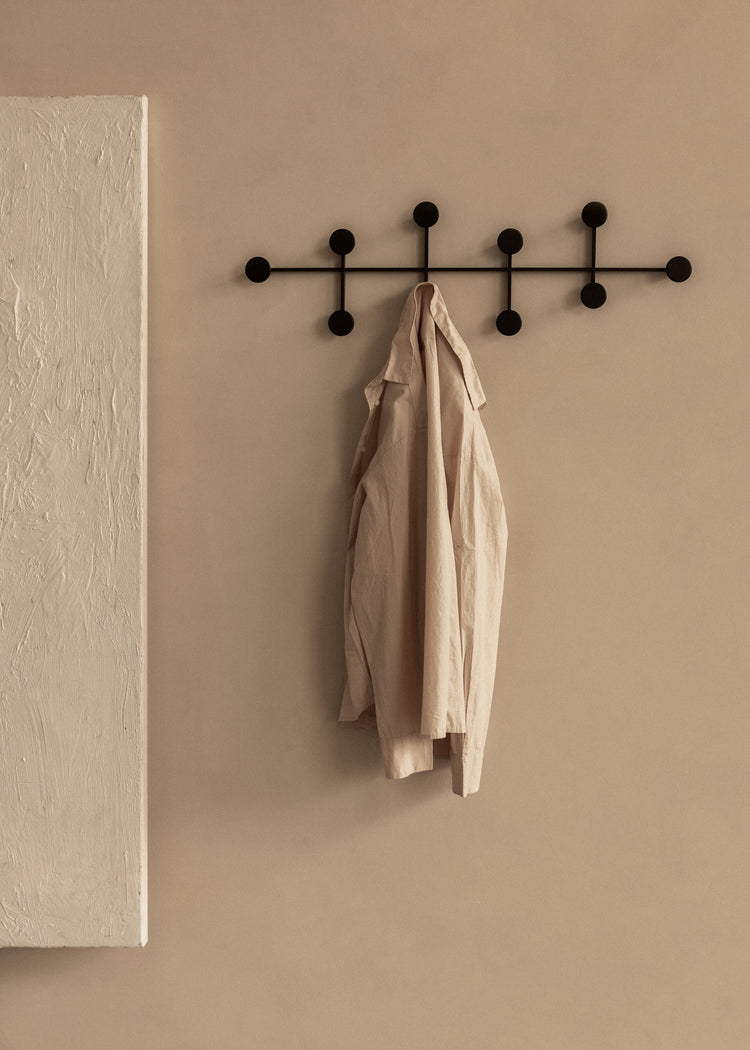 Black metal wall coat rack with round pegs holding a beige linen shirt against a warm taupe wall
