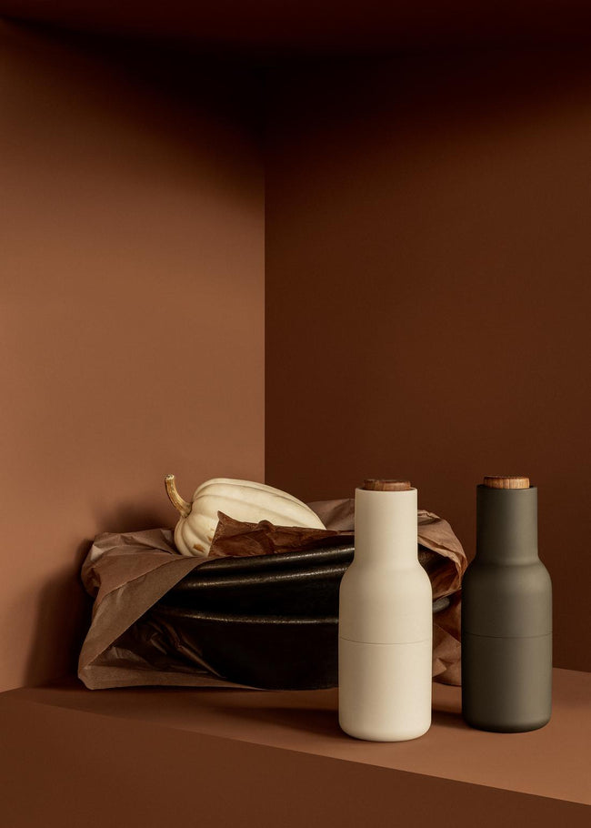 Two matte ceramic grinders with wooden tops, one beige one dark gray, displayed next to a black bowl with a pale gourd on