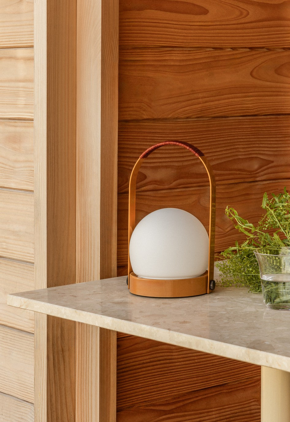Small portable table lamp with white frosted globe, tan leather base and arched leather-wrapped handle on marble shelf.