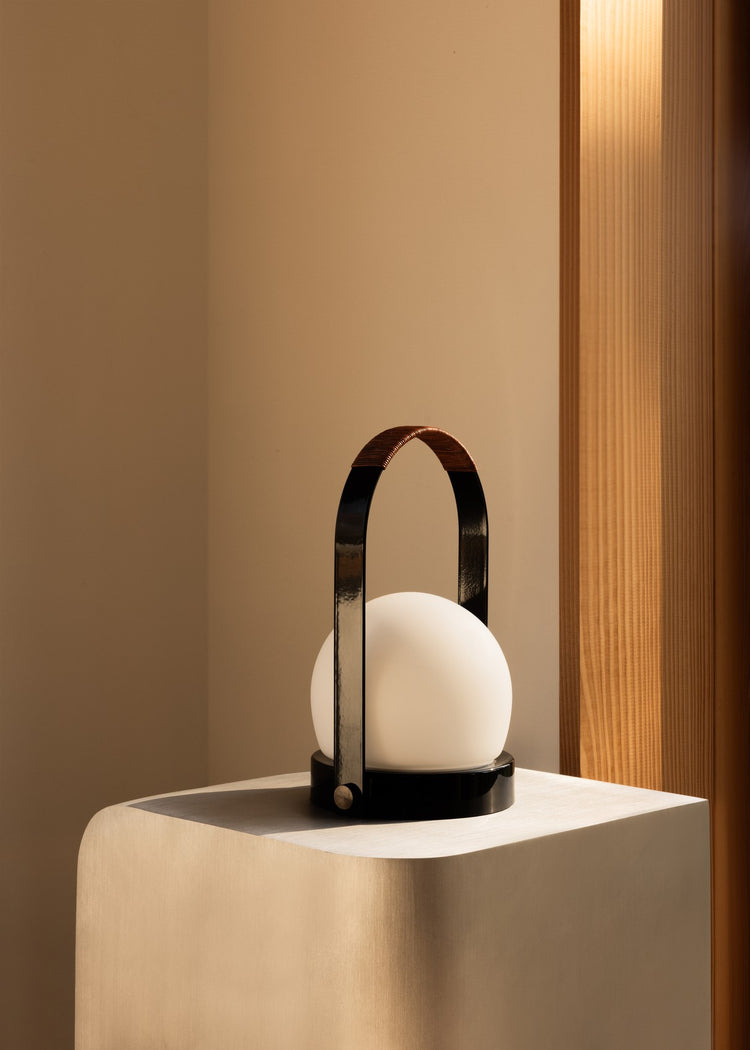 Black metal and leather-handled portable lamp with white frosted glass orb on beige pedestal, warm wood background.