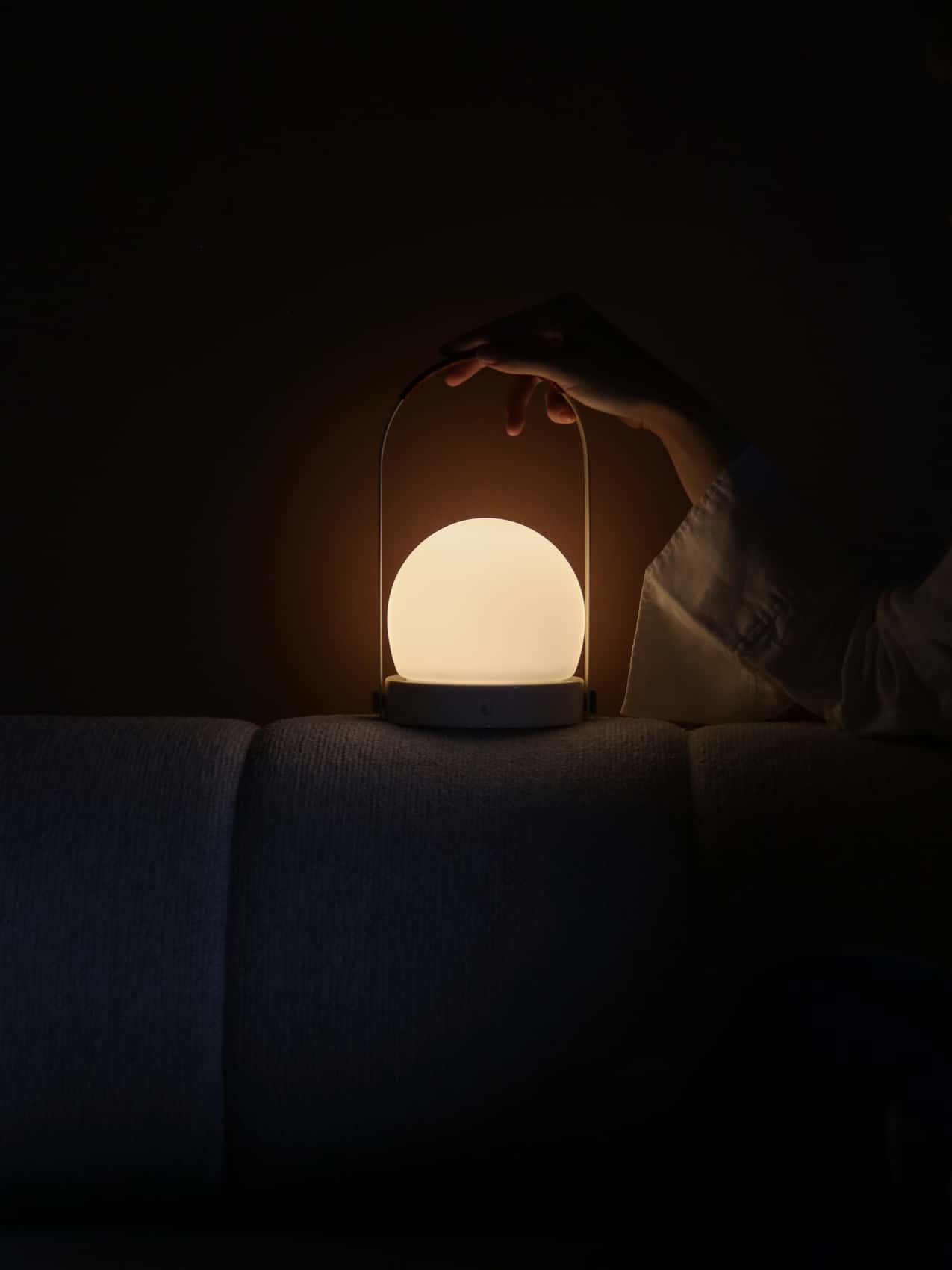 Carrie Portable Lamp with leather handle emitting a soft, warm glow on a textured sofa in a dark interior setting.