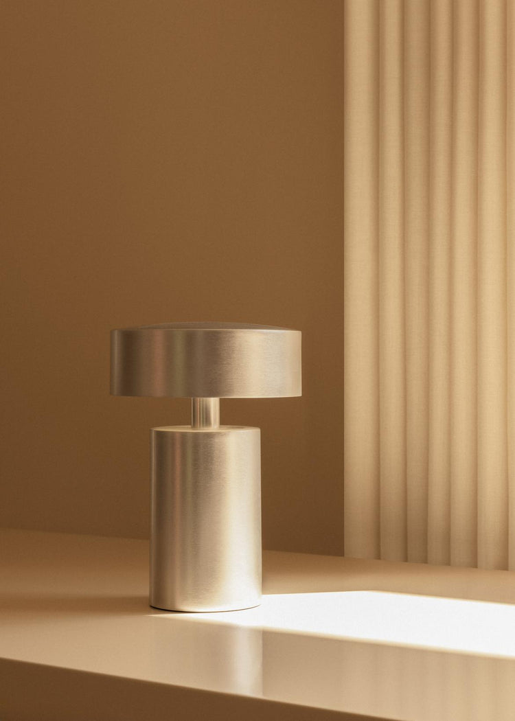 Brushed brass cylindrical table lamp with round disc shade on beige tabletop against warm tan background