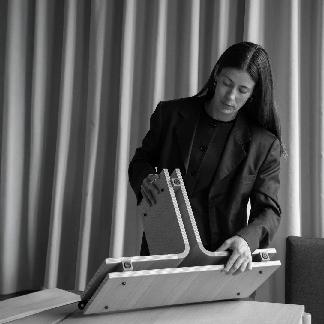 Black-and-white photo of a person assembling a light oak plywood table base with interlocking curved legs and visible screws