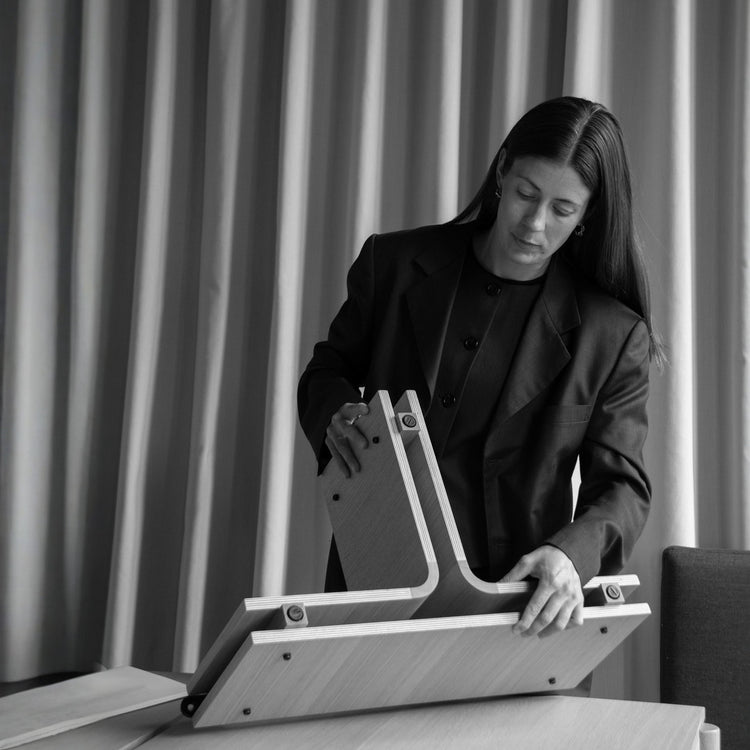 Black-and-white photo of a person assembling a light oak plywood table base with interlocking curved legs and visible screws