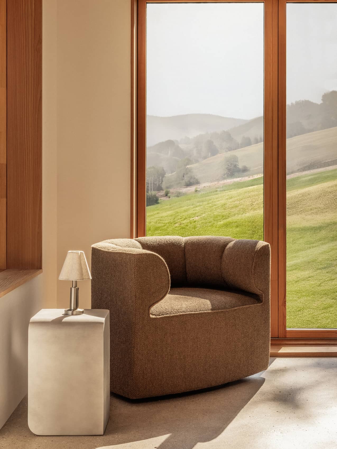 Eave Seamline Lounge Chair in Nutmeg fabric beside a Volume Side Table and Admiral Lamp with a scenic green hill view.