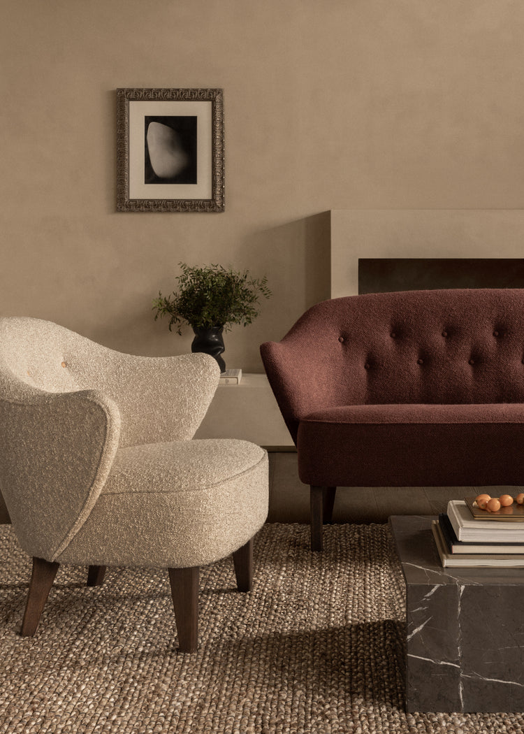 Beige boucle lounge chair and burgundy tufted sofa on woven jute rug, dark wooden legs, marble coffee table and framed wall