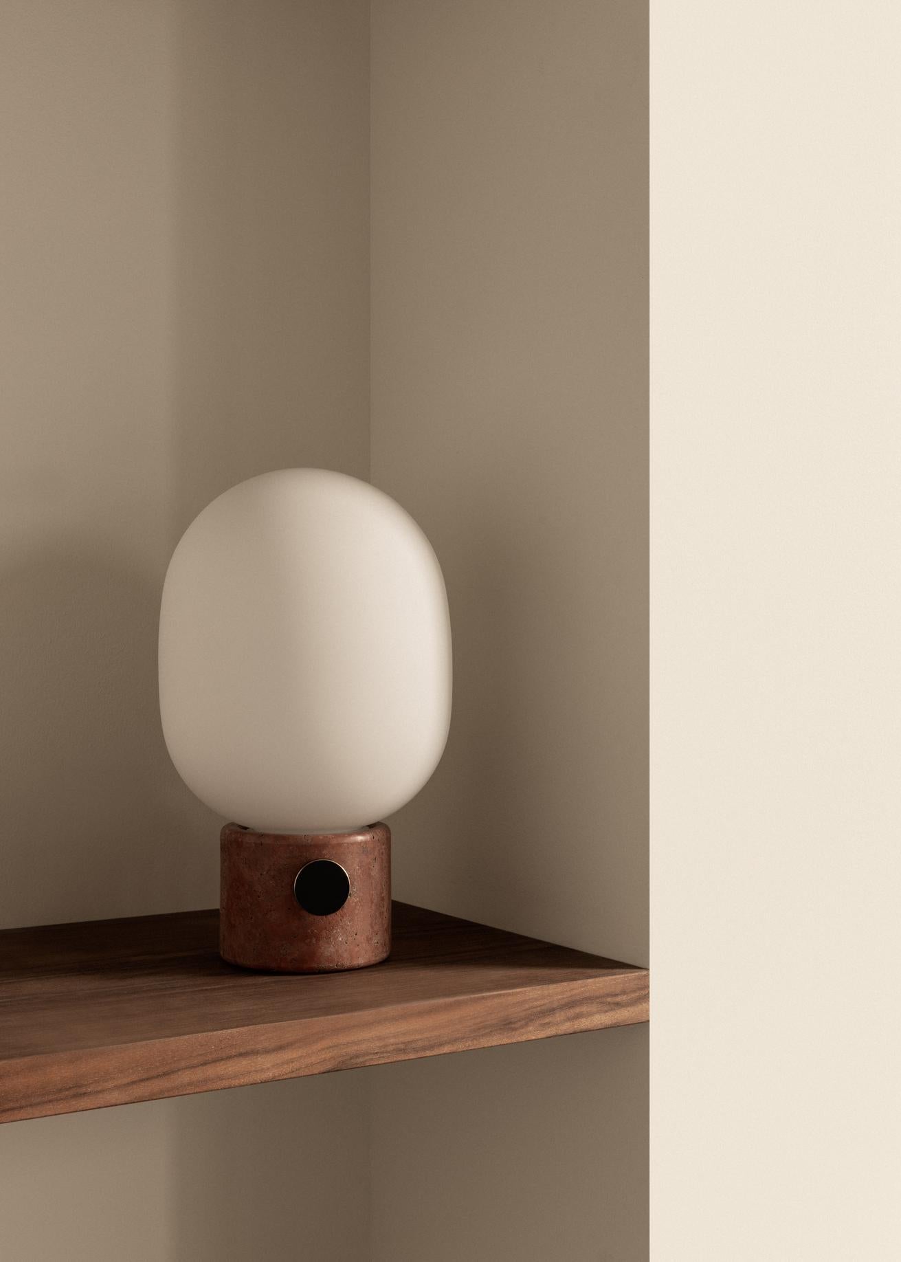 Minimal table lamp with matte white oval glass shade on a cylindrical terracotta base, placed on a wooden shelf.