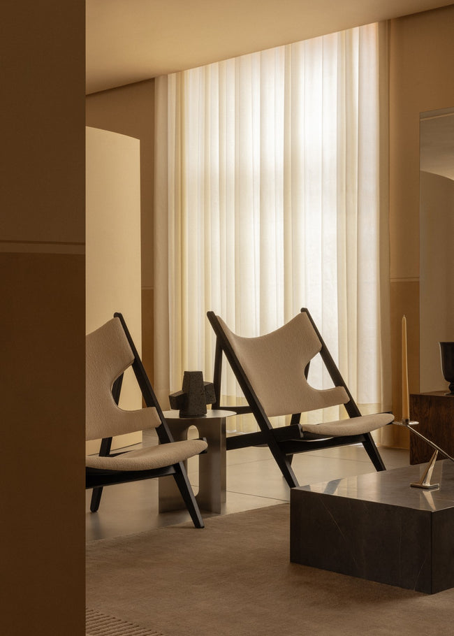Pair of beige upholstered lounge chairs with black wooden frames beside a small metal side table and sheer floor-to-ceiling