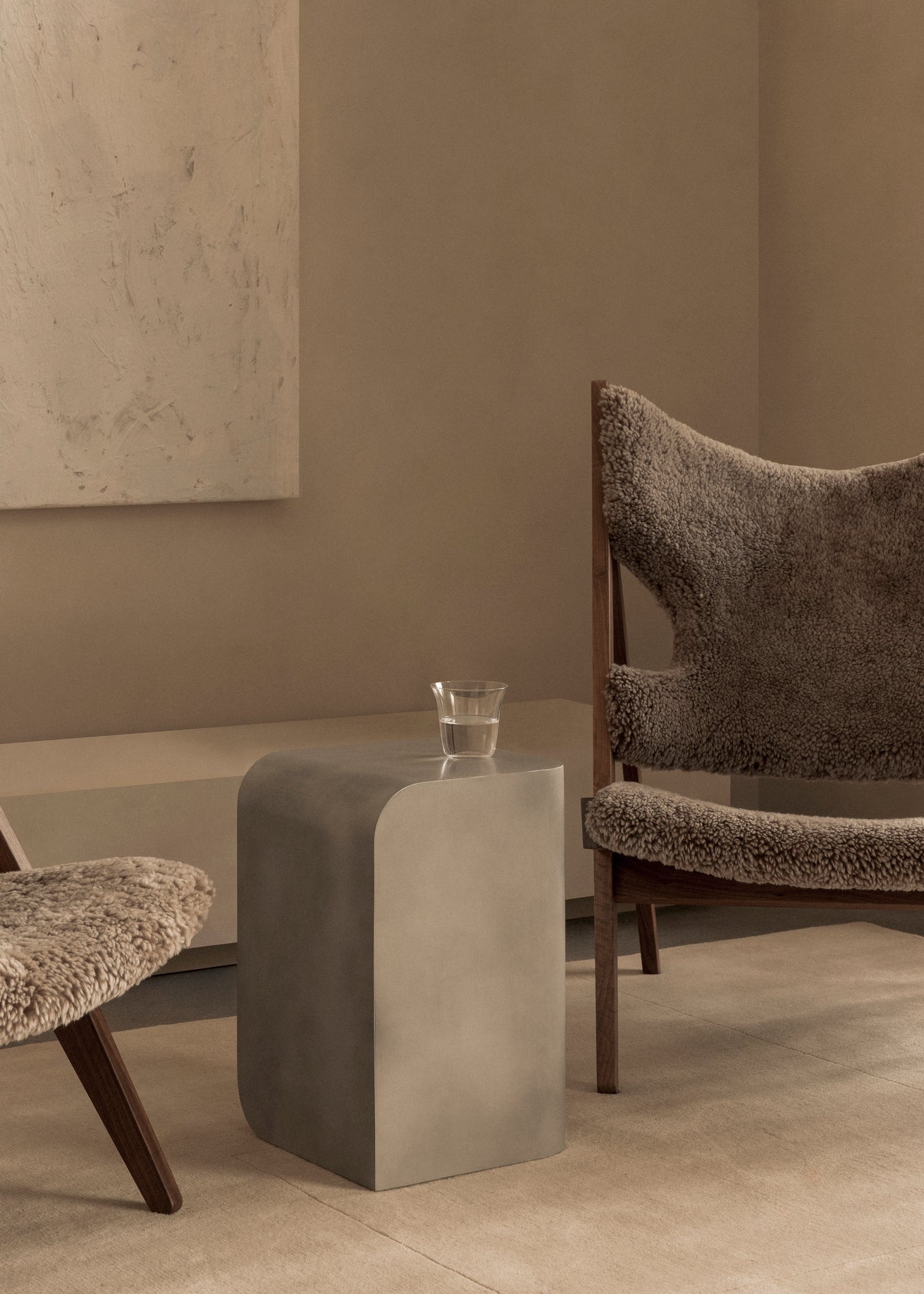Minimal beige living space with a sculpted matte grey side table, glass of water and wool-covered wooden lounge chairs
