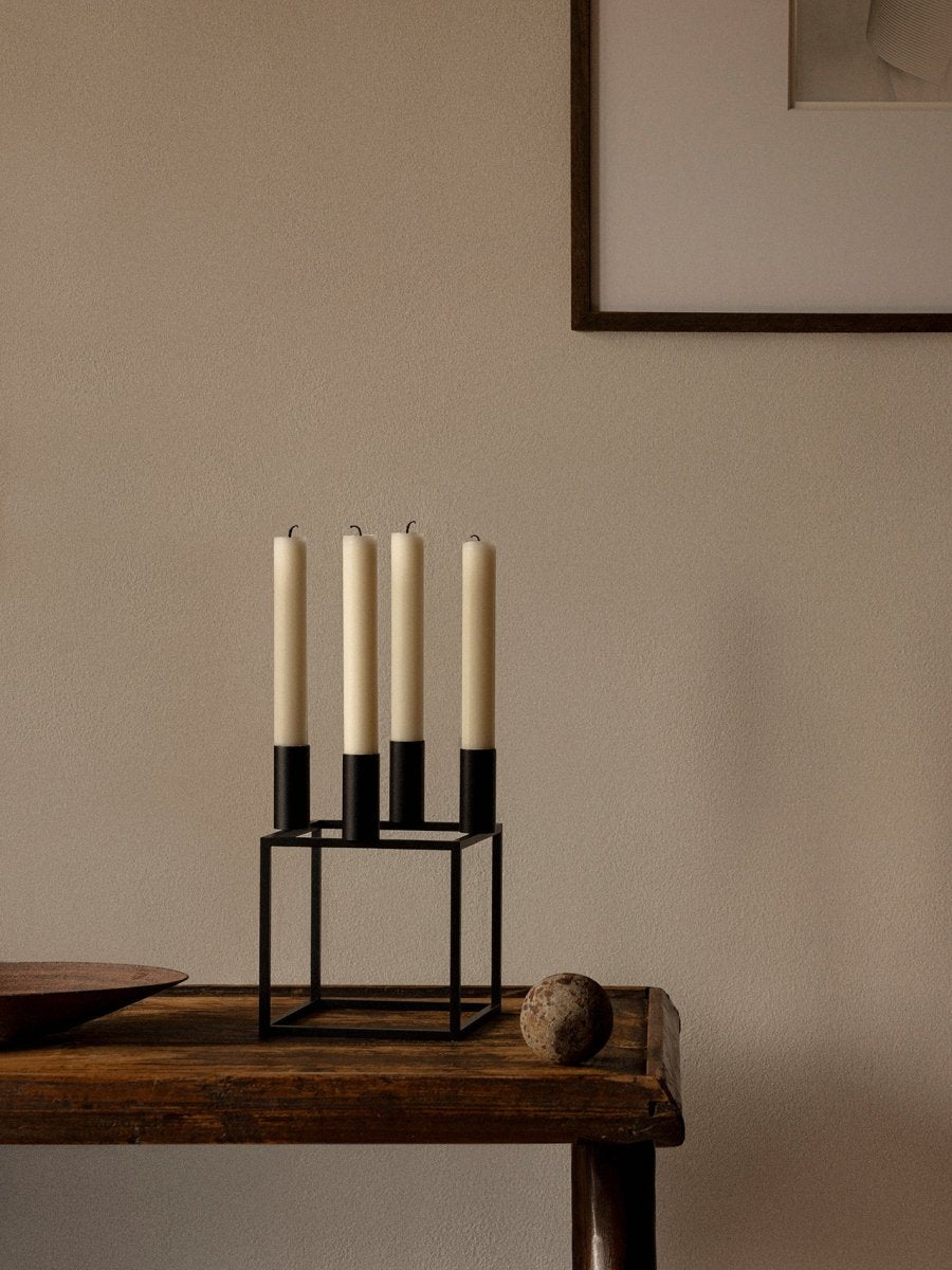 The black Audo Kubus 4 Candle Holder with four tall, off-white candles, styled on a dark wooden table against a neutral wall.