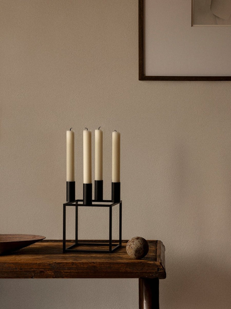 The black Audo Kubus 4 Candle Holder with four tall, off-white candles, styled on a dark wooden table against a neutral wall.