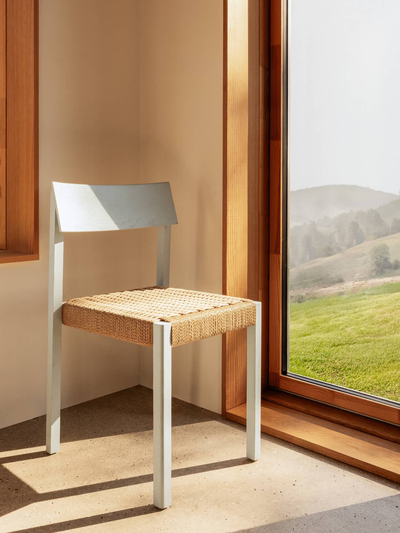 Mauro Dining Chair in Linen Blue Oak with a woven paper cord seat, standing in a sunlit corner by a window.