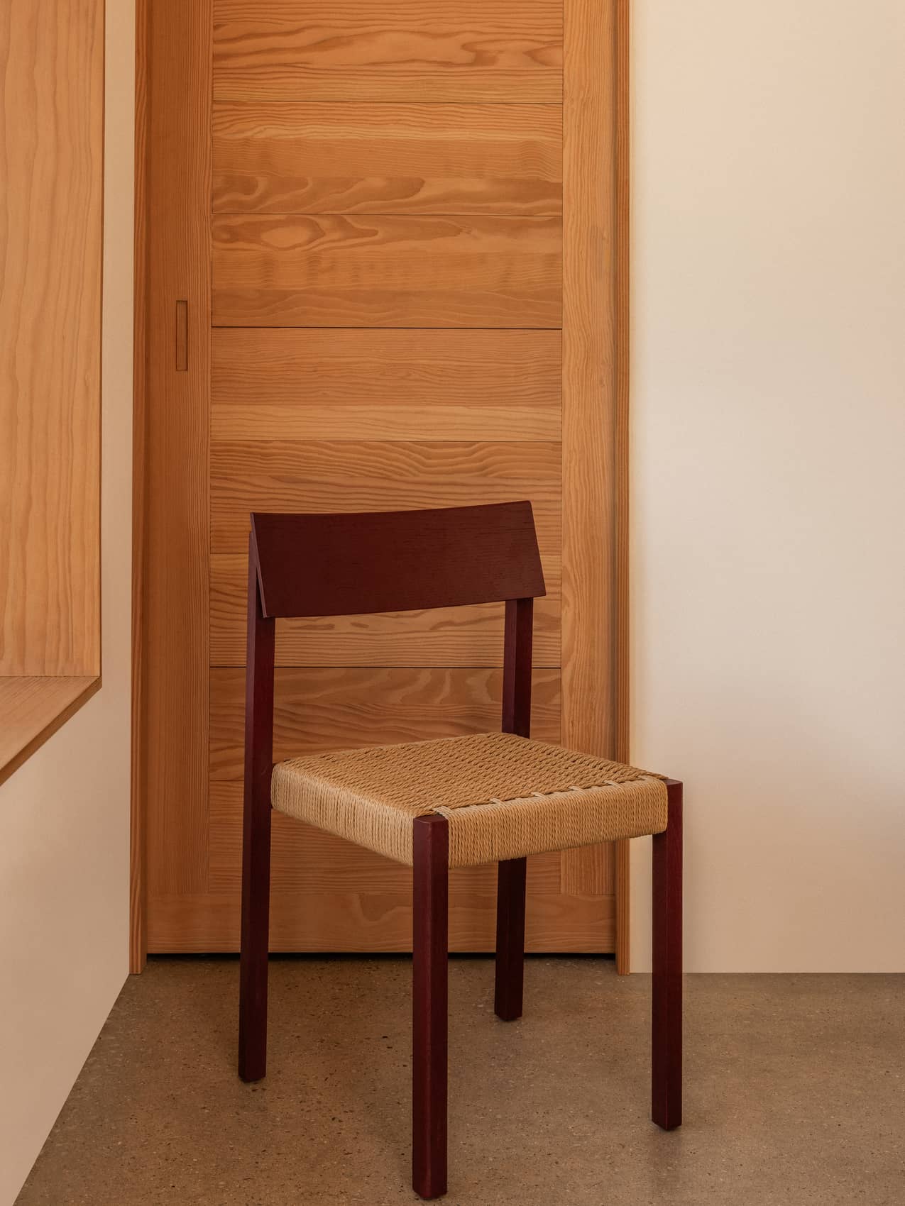 Mauro Dining Chair in French red oak with a handwoven paper cord seat, set against a minimalist wooden door.