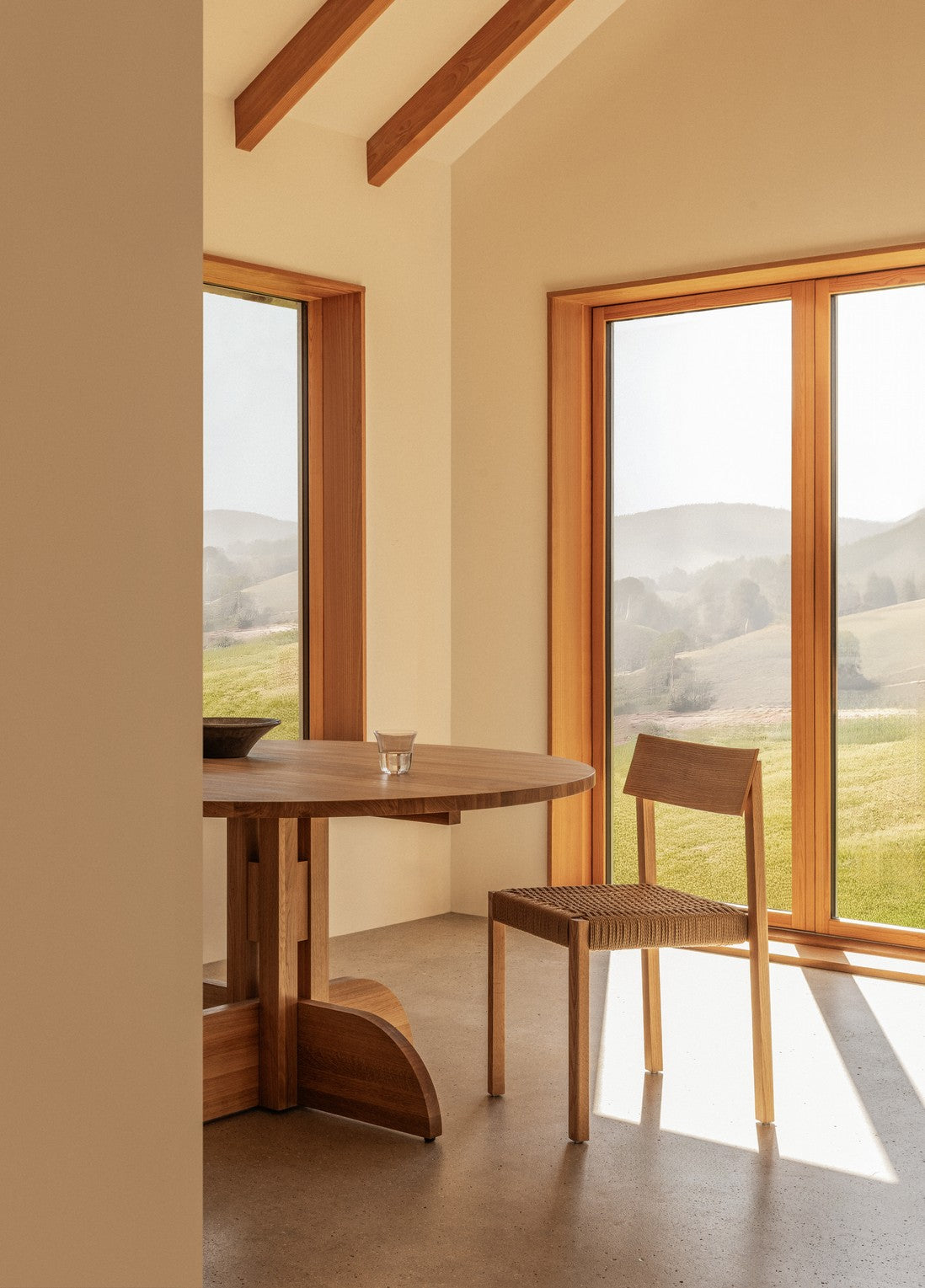 Light oak round dining table and woven-seat wooden chair beside large windows with soft countryside view.
