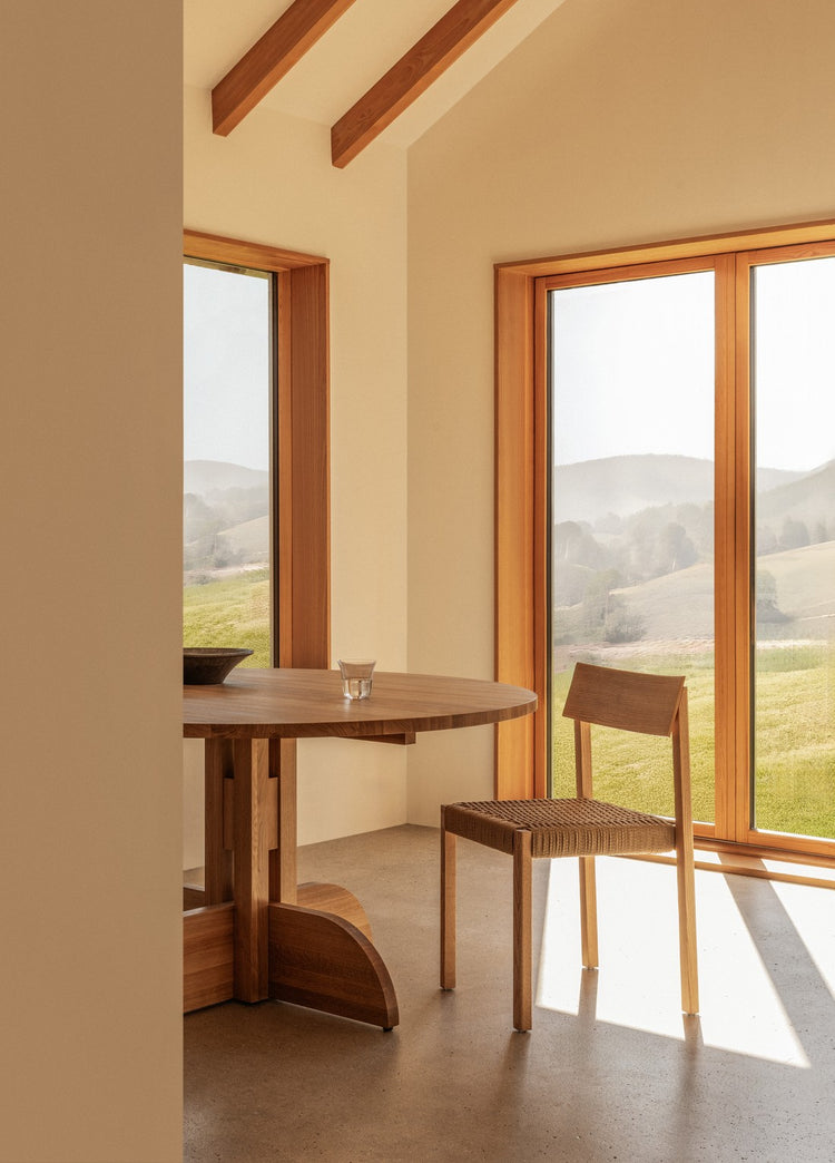 Light oak round dining table and woven-seat wooden chair beside large windows with soft countryside view.