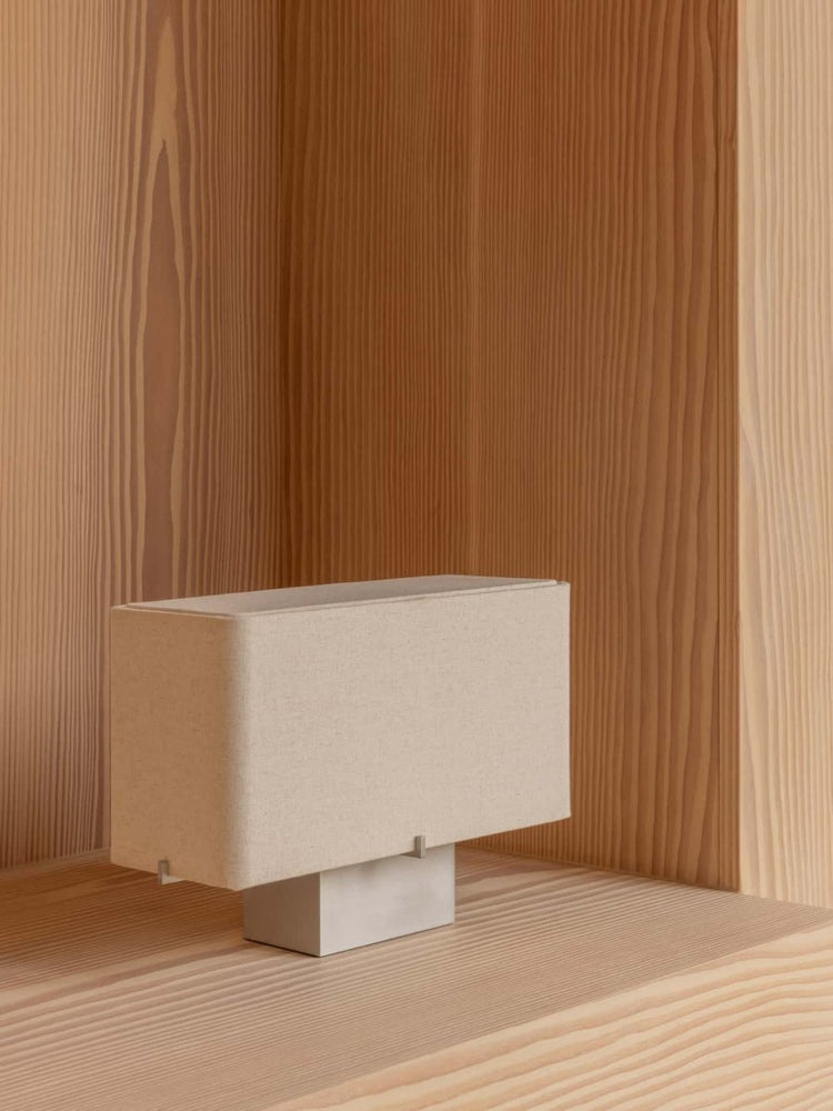 The Nonna Table Lamp, with its rectangular beige textile shade and brushed aluminum base, resting in a light wood alcove.