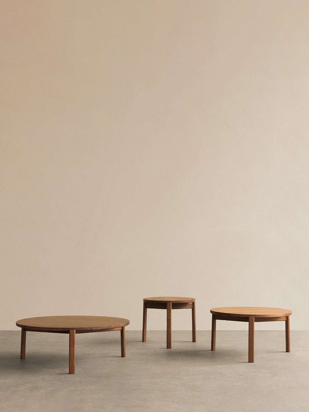 Three round oak side tables of varying heights with tapered legs on a grey concrete floor against a beige wall