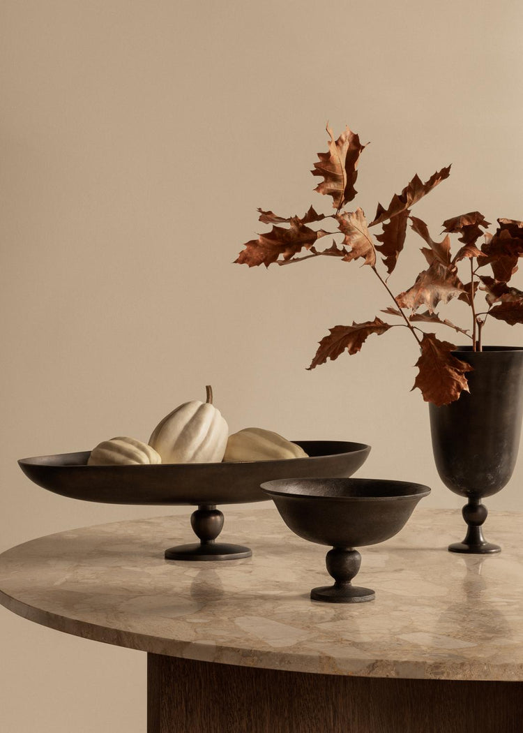 Dark bronze pedestal bowls and tall vase on marble table, with pale decorative squash in a shallow bowl and dried brown oak