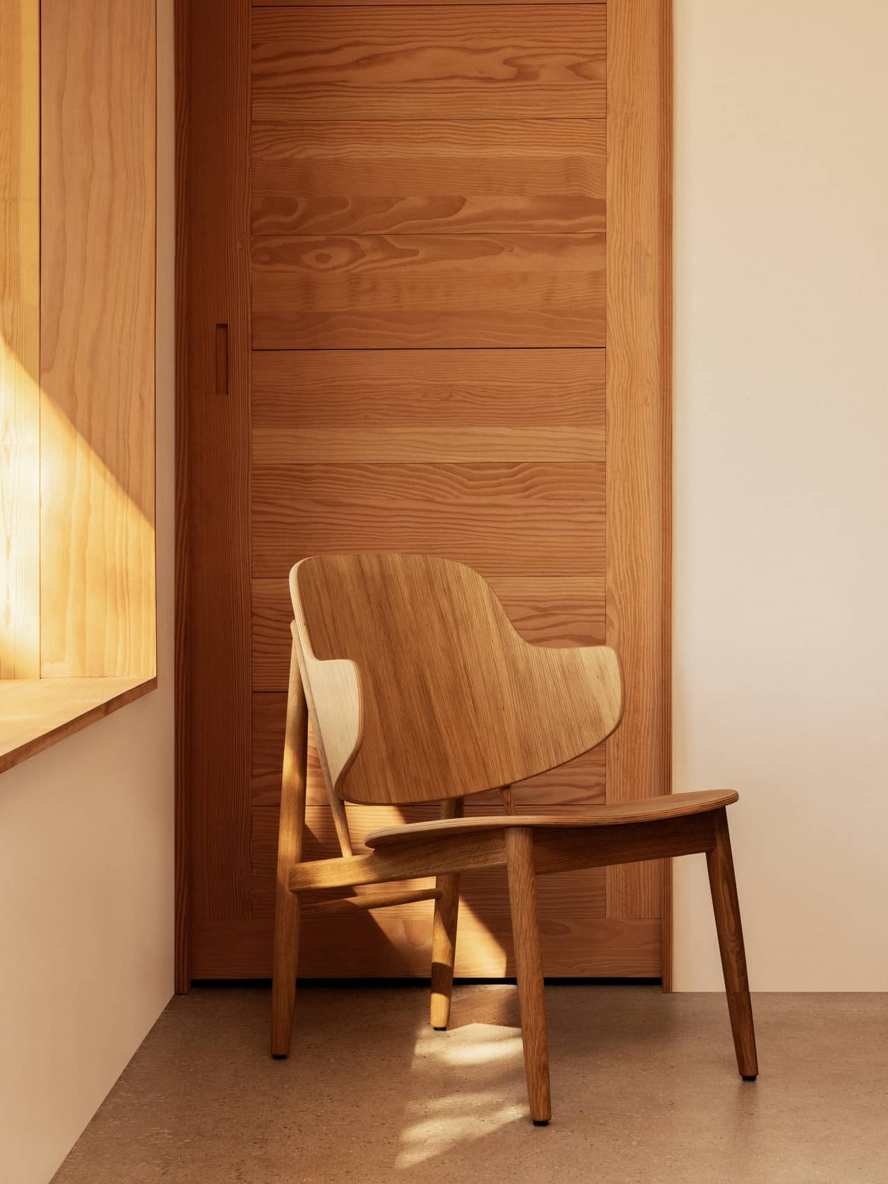 The Penguin Wooden Lounge Chair in natural oak, positioned in a sunlit room against a matching wooden door.