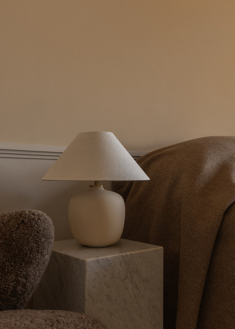 Beige ceramic table lamp with wide fabric shade on a marble cube, next to textured brown throw and boucle chair.
