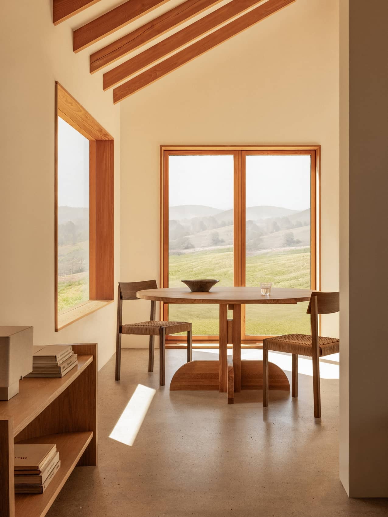 Oak Puffin Dining Table and Mauro Dining Chairs in a sunlit room with Crescent Shelving and views of rolling green hills.