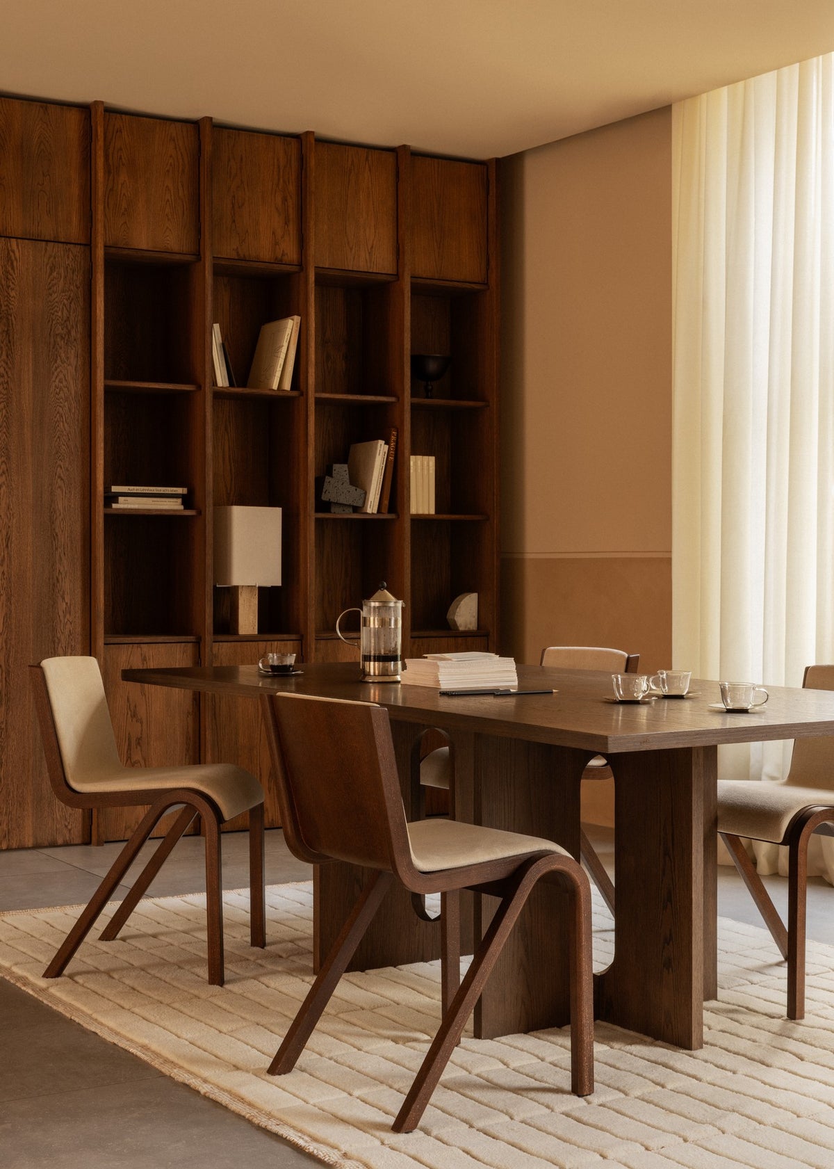 Minimalist dining table setting with dark oak table and curved wood-and-beige upholstered chairs, bookshelf backdrop, coffee