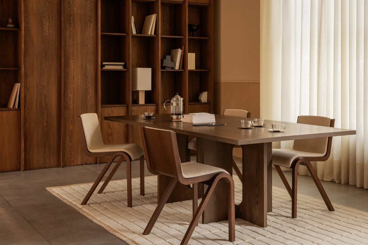 Minimalist dining room with dark walnut rectangular table, curved wooden chairs with beige upholstery, rug and shelving