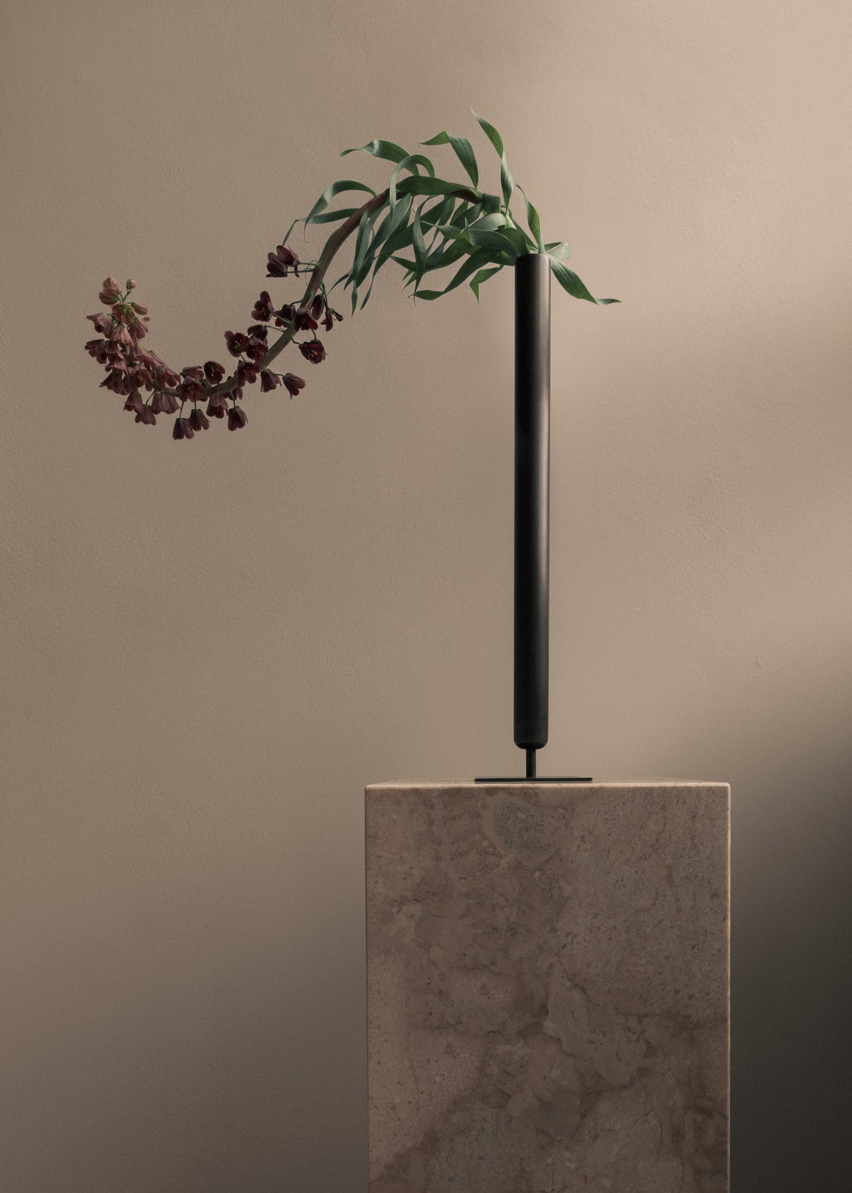 Tall matte black cylindrical metal vase on a stone pedestal holding a drooping stem with green leaves and dark burgundy