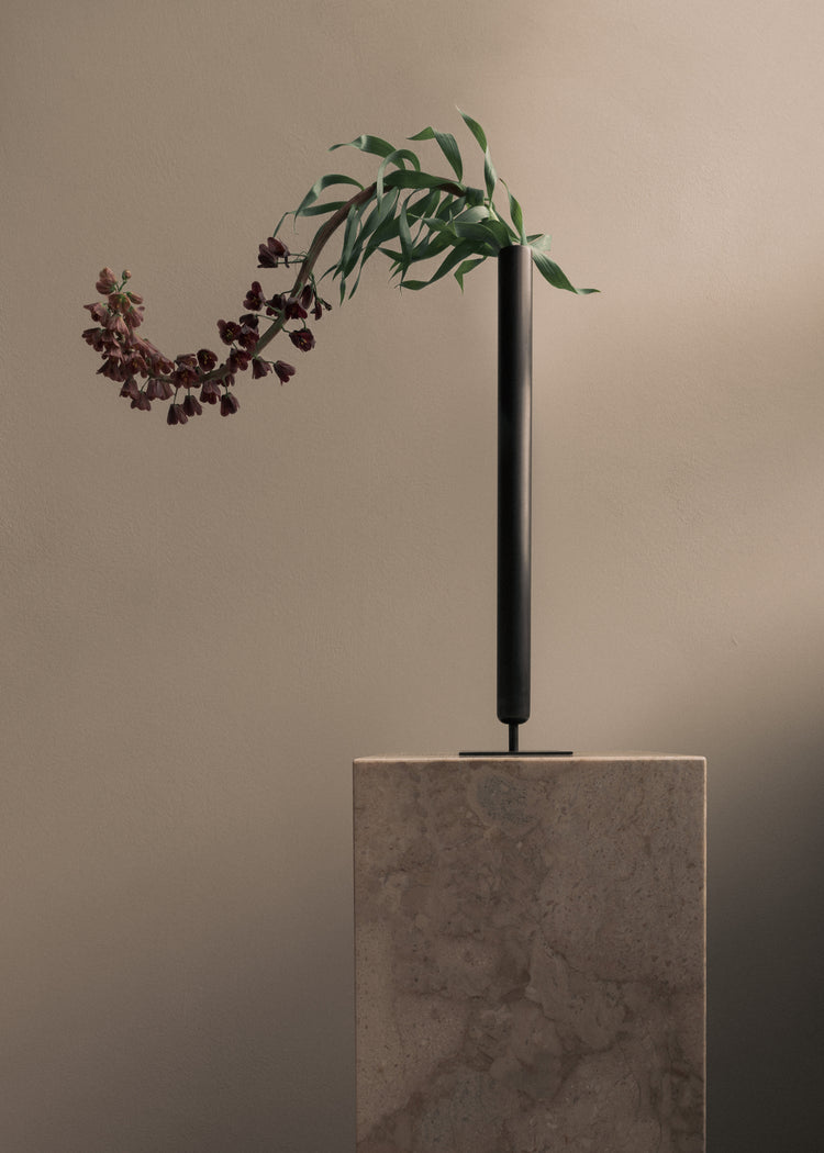 Tall matte black cylindrical metal vase on a stone pedestal holding a drooping stem with green leaves and dark burgundy
