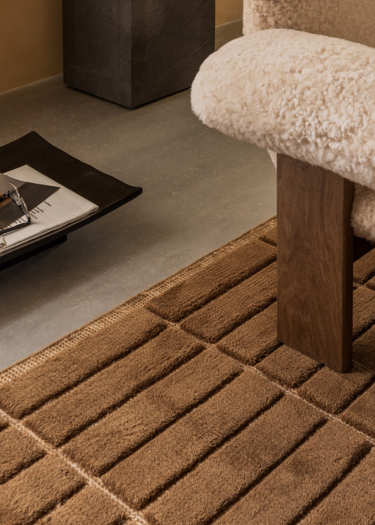 Close-up of brown textured wool rug and wooden bench leg with cream sheepskin seat and black tray holding papers.
