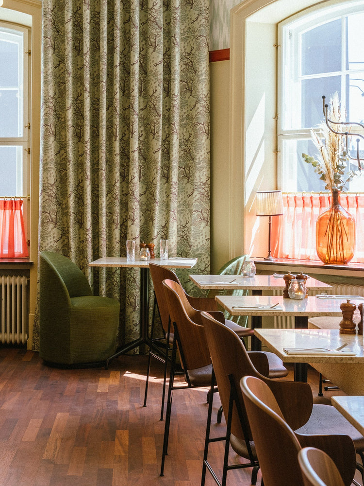 Cozy café corner with green patterned drapes, wooden chairs, marble tables and an amber glass vase by a sunlit window