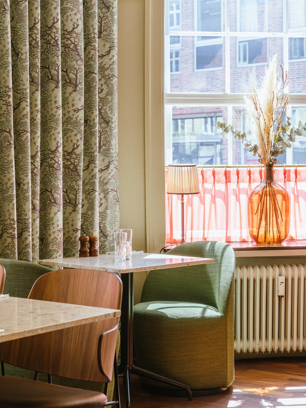 Dining corner with green upholstered chair, marble table, wooden chairs, patterned green curtains and amber glass vase on